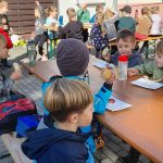 Dzieci siedzą przy stołach piknikowych na świeżym powietrzu, jedzą przekąski i rozmawiają w promieniach słońca.