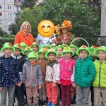 Grupa uśmiechniętych dzieci w zielonych kapeluszach ozdobionych liśćmi, stojących na zewnątrz w towarzystwie opiekunów podczas jesiennego spaceru lub wydarzenia.