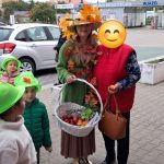 Grupa dzieci w zielonych kapeluszach i dorośli w jesiennych strojach na parkingu, z kobietą trzymającą kosz jabłek pośrodku.