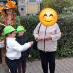 Dzieci w zielonych kapeluszach i opiekunka w jesiennej stylizacji wręczają upominek przechodniowi na osiedlowym chodniku.