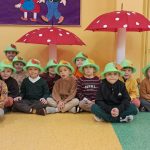 Grupa przedszkolaków w zielonych kapeluszach z liśćmi siedzi na podłodze w sali pod dwoma dużymi dekoracyjnymi muchomorami.