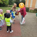 Dziecko w odblaskowej kamizelce wręcza kwiaty kobiecie na chodniku w obecności grupy przedszkolaków i opiekunki.