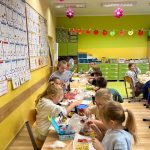 Dzieci w kolorowej klasie szkolnej siedzą przy połączonych ławkach i jedzą przygotowane wcześniej owocowe przekąski oraz szaszłyki.