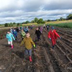 Grupa dzieci w kolorowych kurtkach i butach idzie przez pole pod zachmurzonym niebem, a w tle widać zielone drzewa i uprawy.