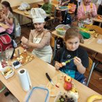Grupa dzieci w wieku szkolnym, ubranych w fartuszki ochronne, przygotowuje zdrowe przekąski na stole zastawionym różnymi owocami, takimi jak jabłka i winogrona, w jasnej sali lekcyjnej.
