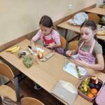 Dwie dziewczynki w fartuszkach siedzą przy szkolnej ławce i robią owocowe szaszłyki ze świeżych owoców.