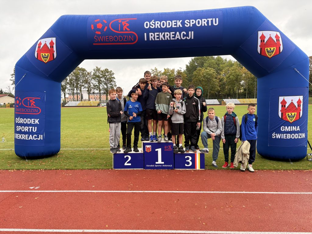 Grupa chłopców stoi na podium dla zwycięzców i wokół niego pod niebieską dmuchaną bramą na boisku sportowym.