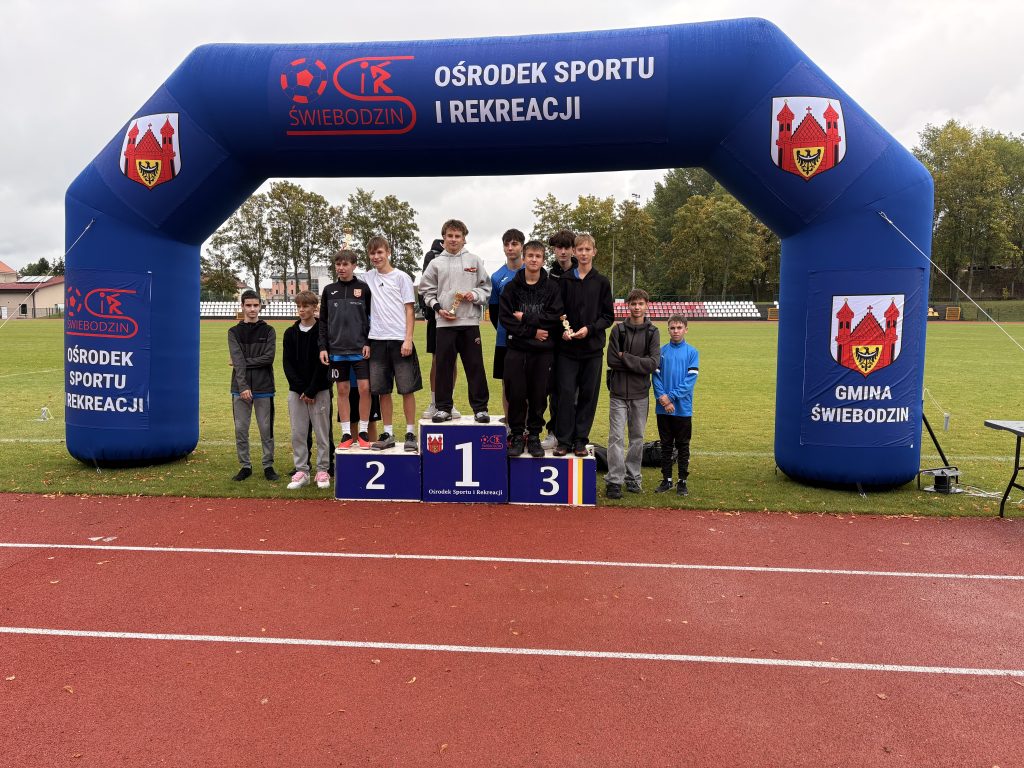 Grupa chłopców stoi na podium dla zwycięzców i wokół niego, pod niebieską dmuchaną bramą na zewnętrznym torze sportowym, pozując do zdjęcia po zakończeniu zawodów sportowych.