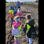 Grupa małych dzieci, ubranych w plecaki i neonowe kamizelki odblaskowe, idzie w szeregu po polnej ścieżce na świeżym powietrzu w towarzystwie dorosłych, pod błękitnym niebem z rozrzuconymi chmurami.
